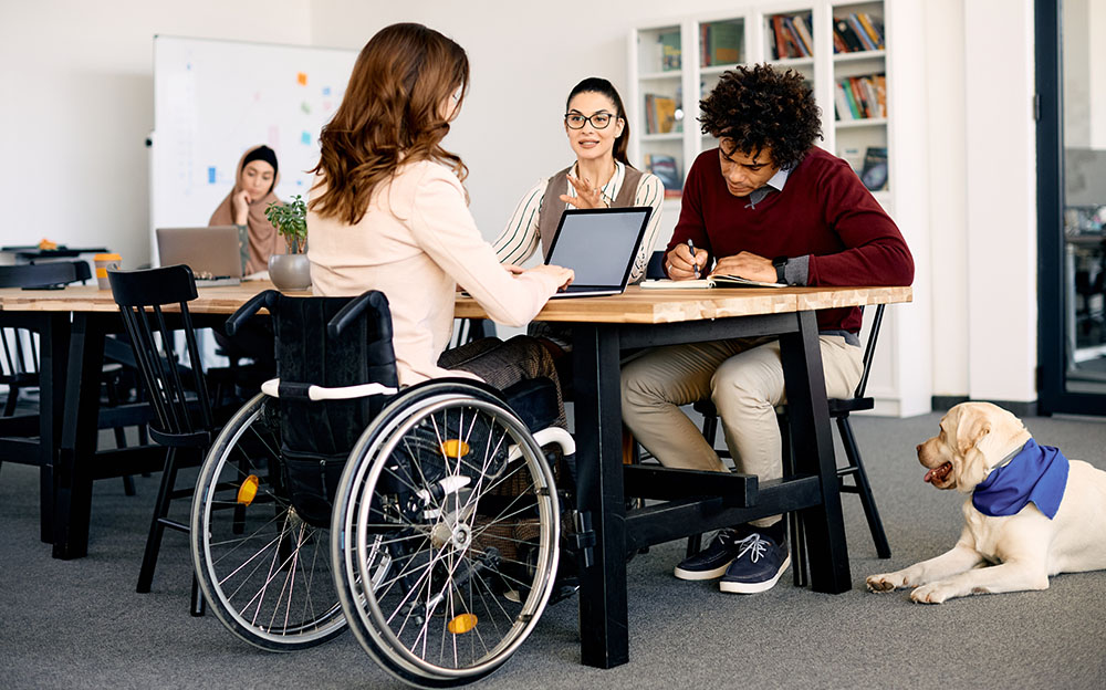 A person in a wheelchair and two other people show inclusion in the workplace as they collaborate at a table next to a service dog.