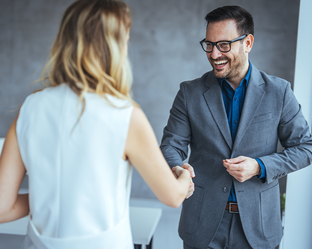 An employer in a white blouse shakes the hand of another person in a gray suit after hiring a Deaf employee.