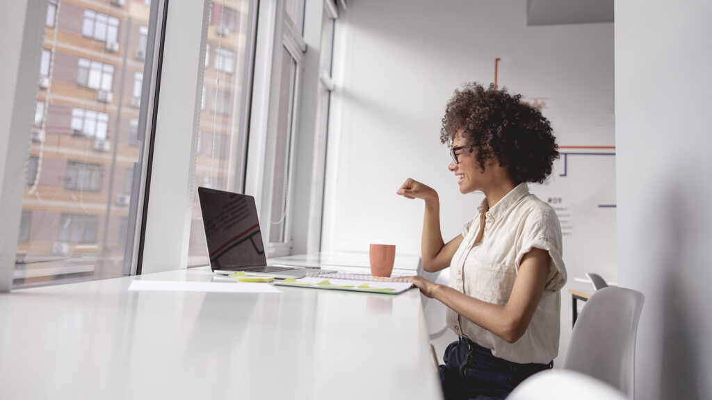 A white room with a desk and big windows. A woman sits in front of a laptop computer and a pink coffee cup, using American Sign Language.