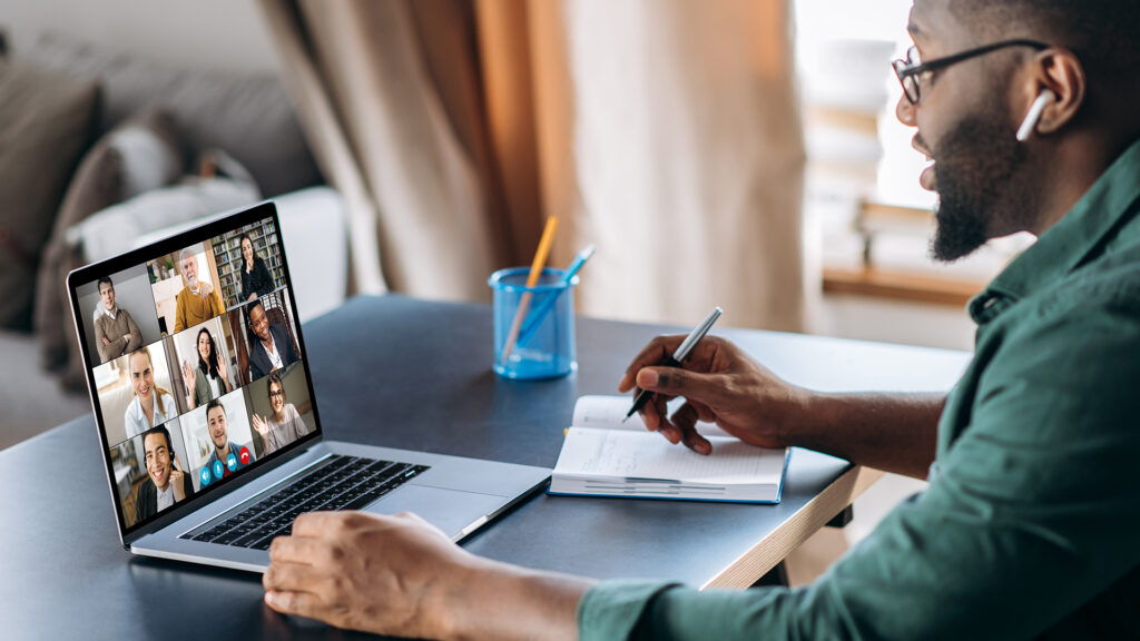 A black man with glasses and an airpod with a green shirt sitting in front of a laptop that features people in a virtual meeting.