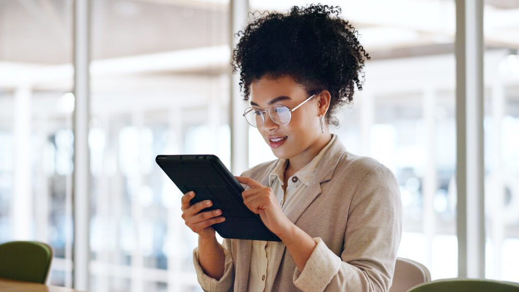 A woman looking at a tablet with computer screens behind her and a colleague sitting to her right.