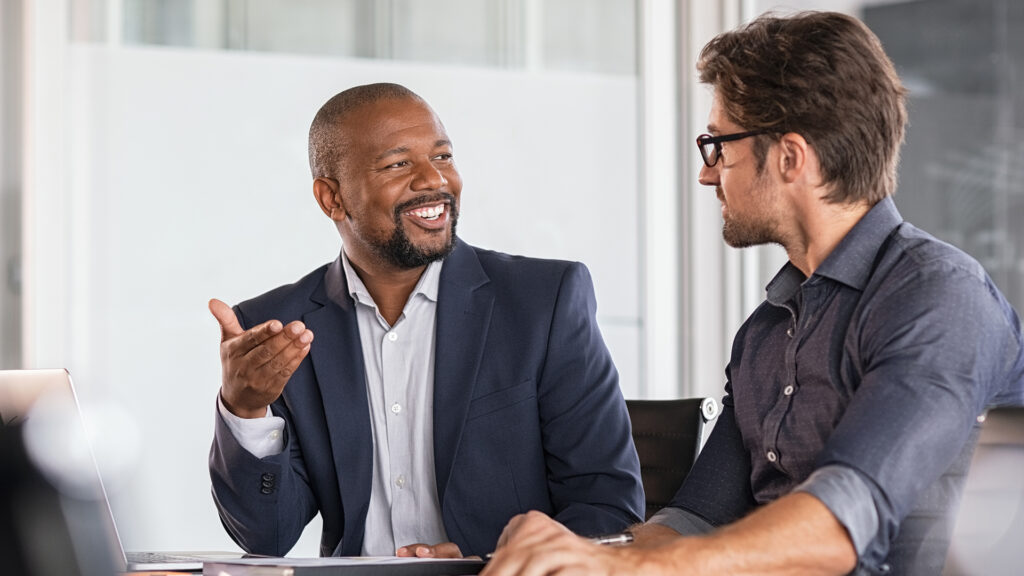 Two men sitting in a conference room having a conversation with a laptop between them.